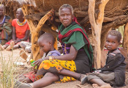 ältere afrikanische Frau sitzt mit zwei kleinen Kindern auf dem Boden, im Hintergrund andere Erwachsene