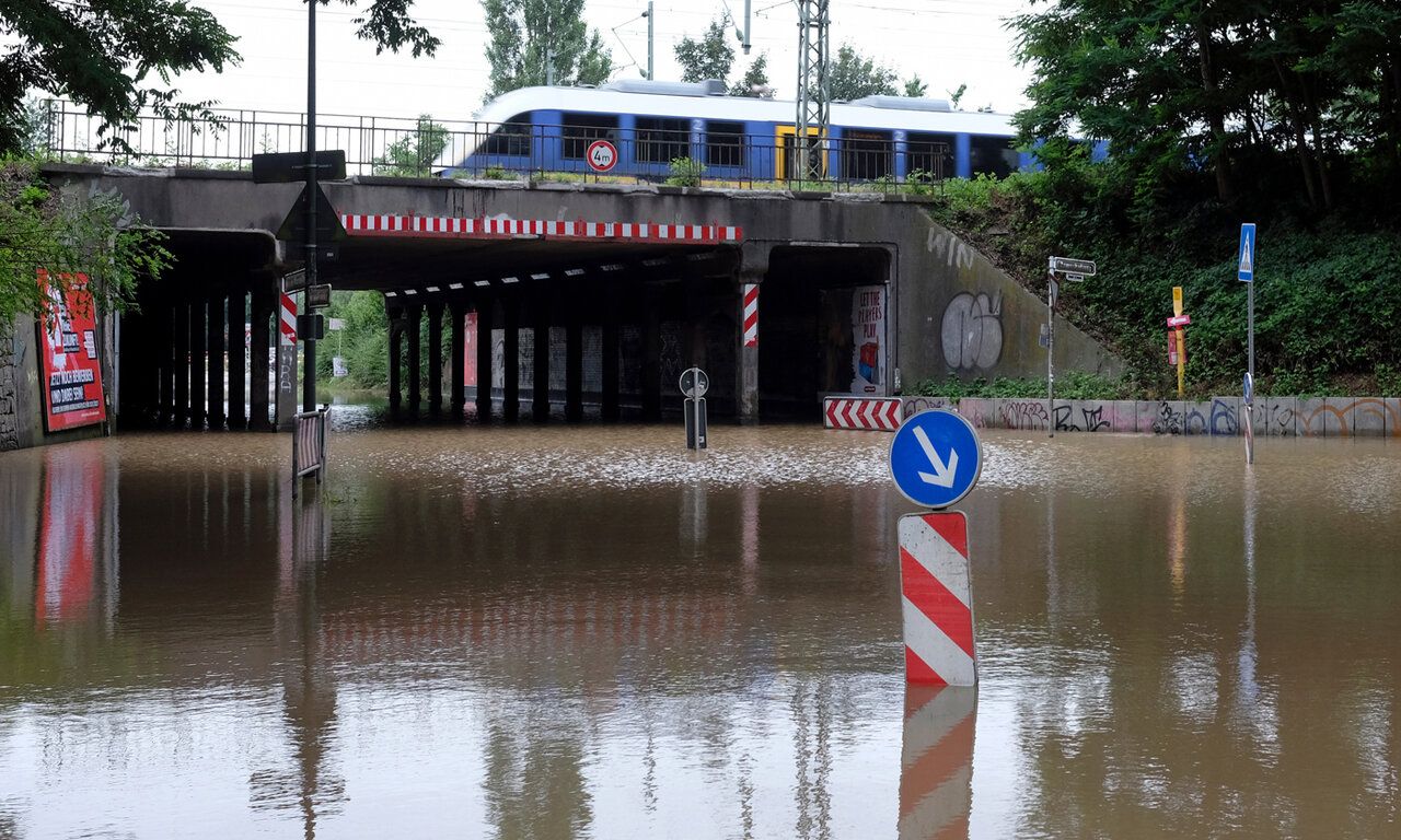 Hochwasser auf einer Straße in Düsseldorf, Straßenschilder stehen im wasser, im Hintergrund eine Unterführung, darauf eine S-Bahn