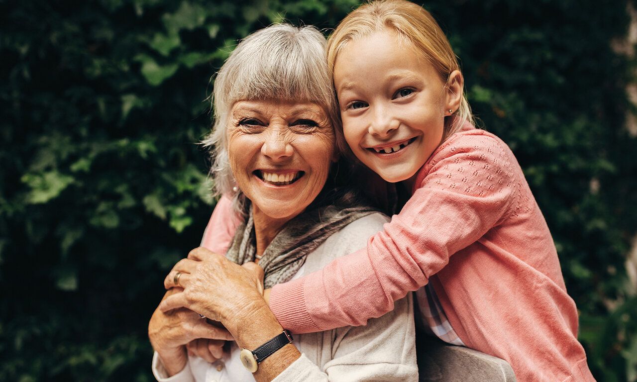 ein Mädchen umarmt seine Oma von hinten, beide lachen in die Kamera