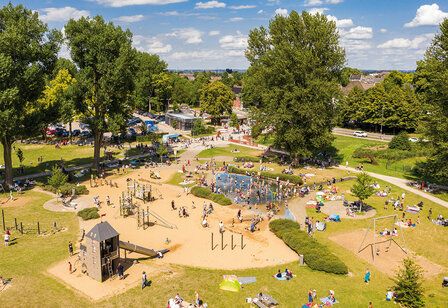 Eine Wiese mit einem Spielplatz und Bäumen von oben, viele Menschen tummeln sich dort