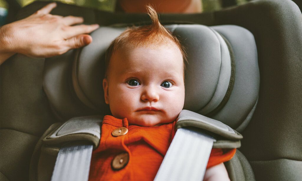 Ein Baby sitzt im Auto in einem Kindersitz, guckt lustig und hat eine Haartolle