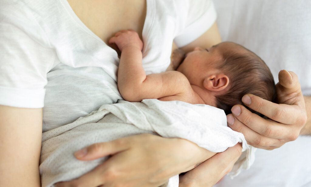 Baby wird von der Mutter im Arm gehalten und gestillt, die Hand des Vaters hält das Köpfchen