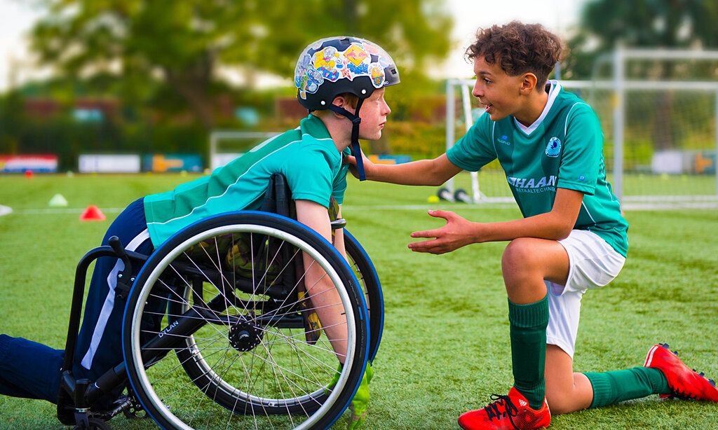 Filmszene aus Bleib am Ball, ein Junge mit Helm kniet in seinem Rollstuhl, ein andere kniet vor ihm und redet auf ihn ein, beide sind auf einem Fußballplatz