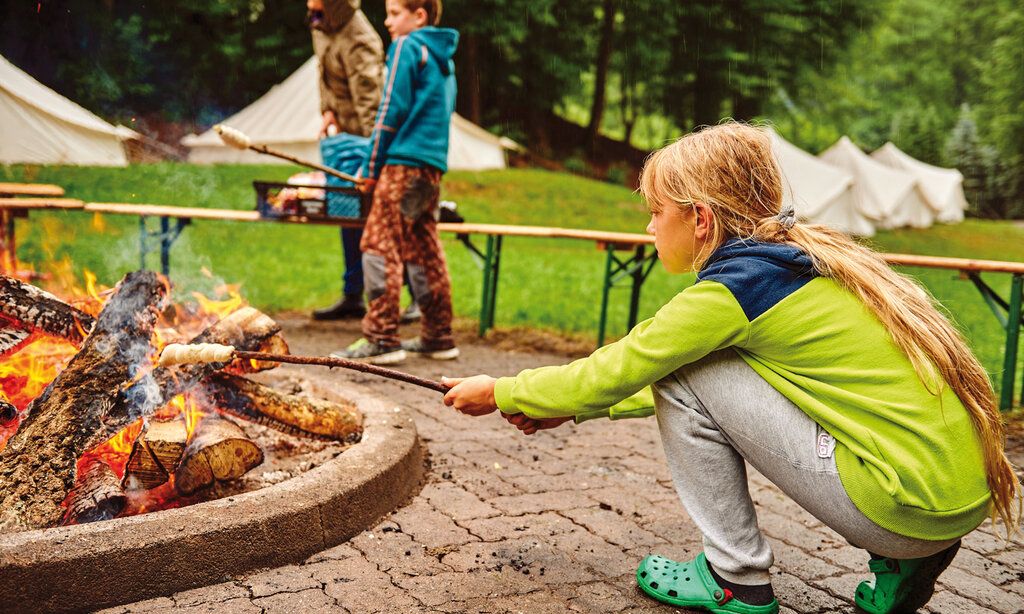 ein Mädchen hält einen Holzstock mit Brotteig ins Lagerfeuer und macht Stockbrot, im Hintergrund zwei Personen und Zelte auf einer Wiese