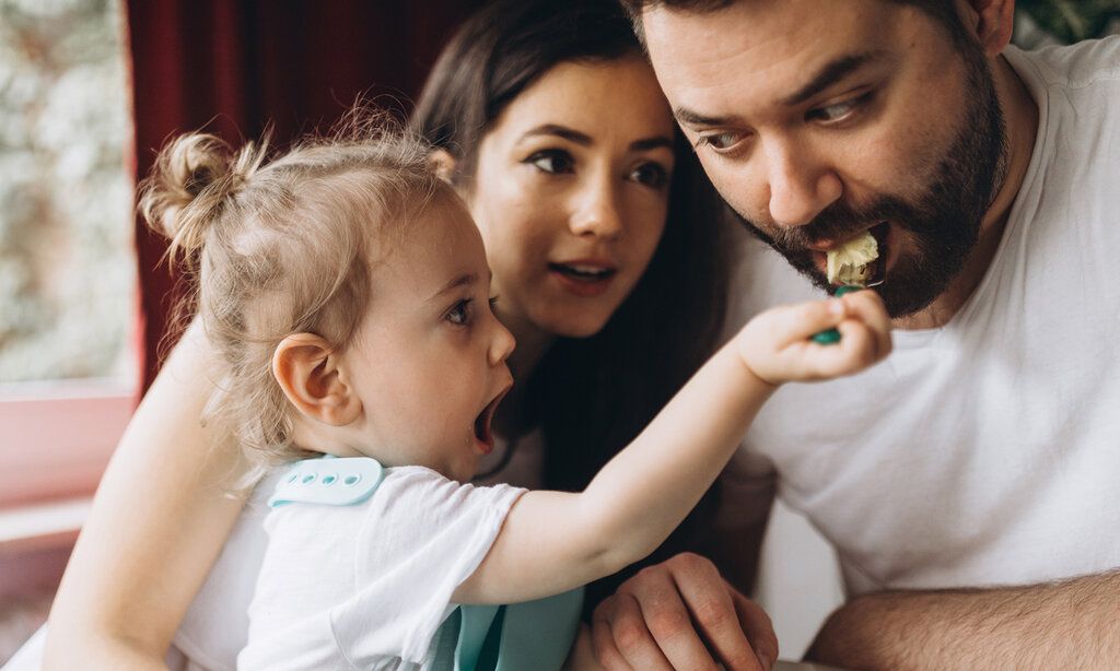 Kind füttert Vater mit einem Löffel, die Mutter guckt zu