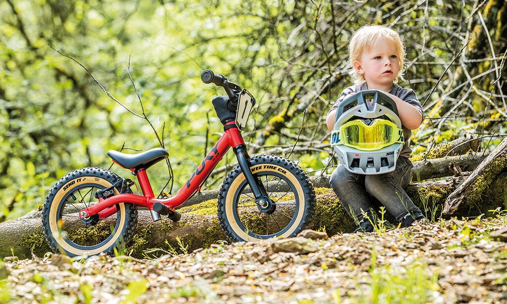 Kleines Kind sitzt auf einem Baumstamm, auf dem Schoß ein Fahrradhelm, neben ihm ein Laufrad