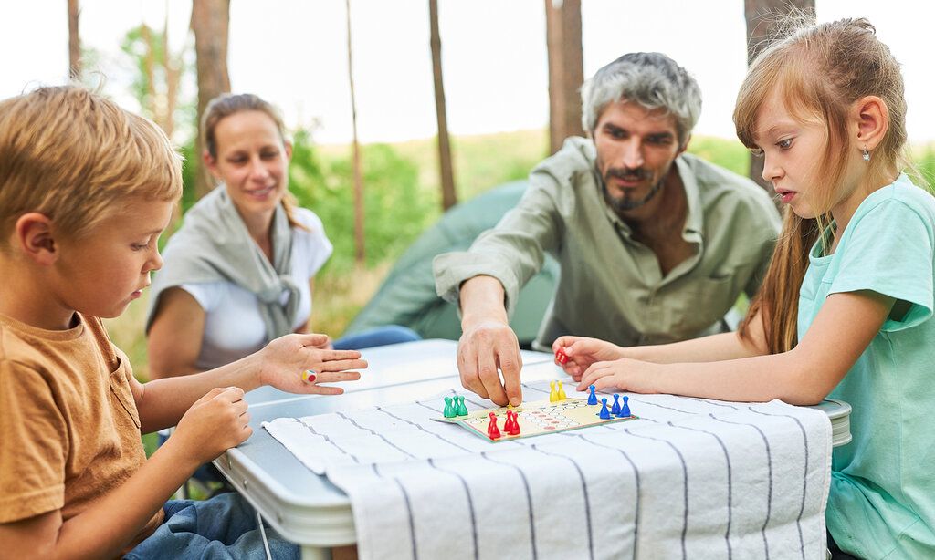 eine Familie spielt Mensch-ärgere-dich-nicht auf einem Campingtisch im Wald