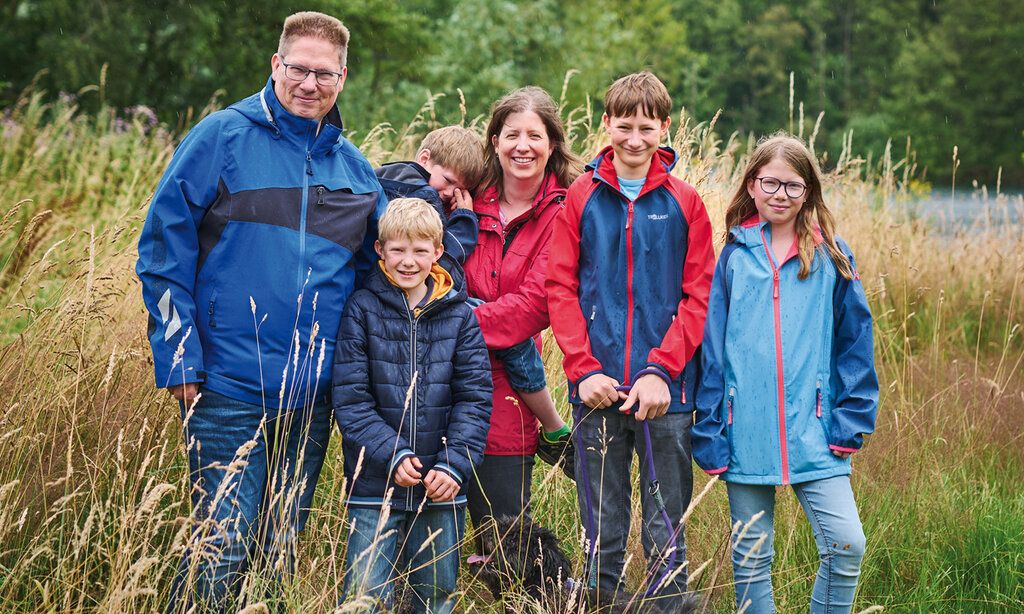 Porträt von familie Teichmann aus Grevenbroich. Vater, Mutter, drei Jungs, ein Mädchen und ein Hund stehen auf einer hochgewachsenen Wiese. Die Mutter trägt den kleinsten Sohn auf dem Arm, der weint.