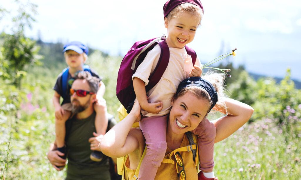 Familie wandert über eine Blumenwiese, Kinder bei Vater und Mutter huckepack