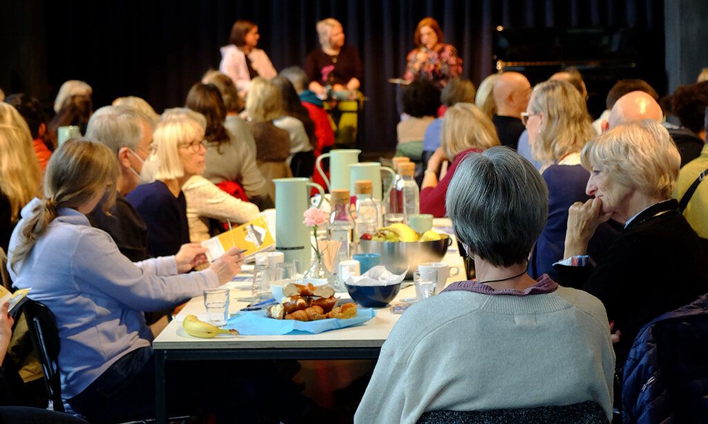 Leute sitzen zusammen an einem Tisch, auf dem Frühstück bereitsteht, im Hintergrund eine Bühne, auf der sich drei Frauen unterhalten