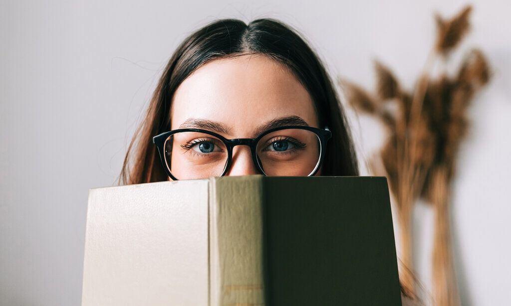 Frau mit Brille hält sich ein Buch vors Gesicht und guckt darüber in die Kamera