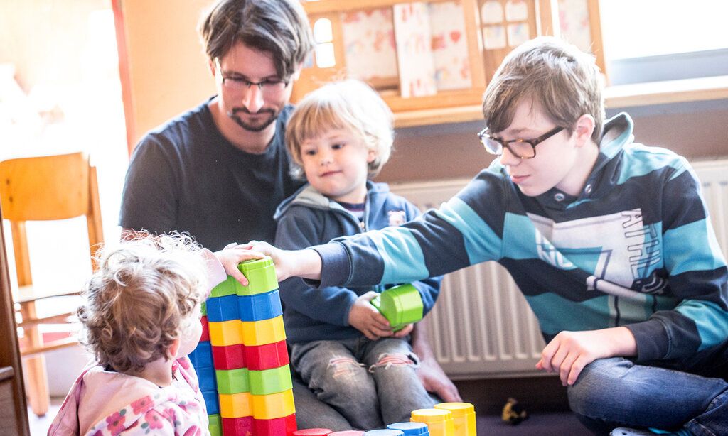 Ein Erzieher und ein Junge sitzen in einer Kita und spielen mit zwei Kleinkindern