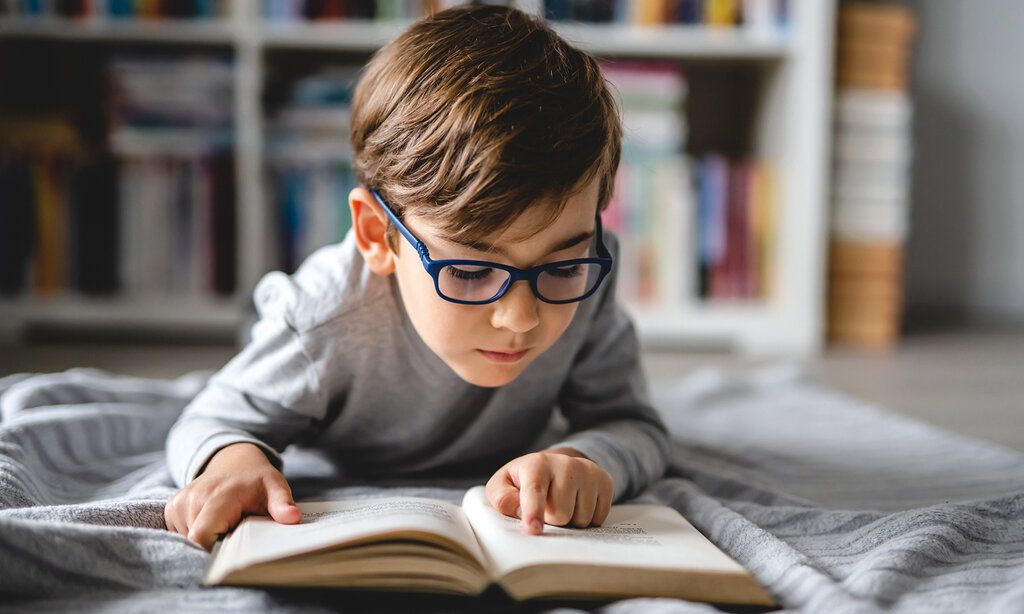 Kleiner Junge mit Brille liegt im Bett undliest ein Buch