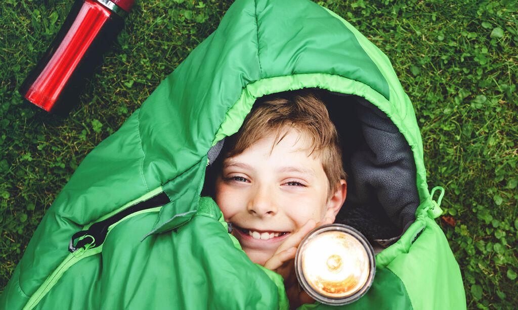 Blick von oben auf einen Junge, der mit Taschenlampe im Schlafsack liegt, Untergrund: Wiese