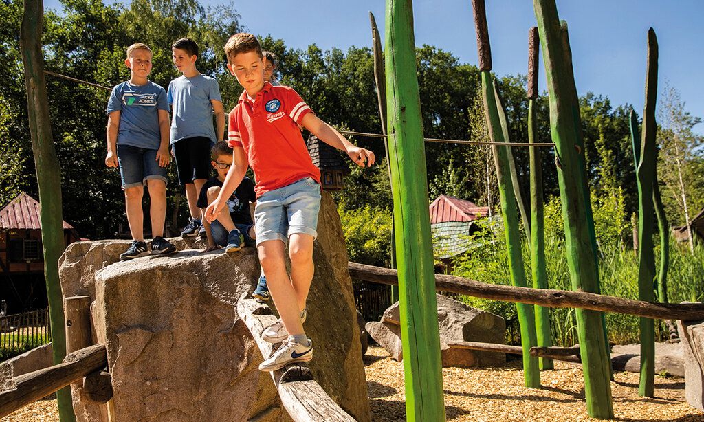 Ein Junge balanciert auf einem Holzbalken auf einem Spielplatz, andere Kinder warten auf einem Felsklotz, dass sie dran kommen