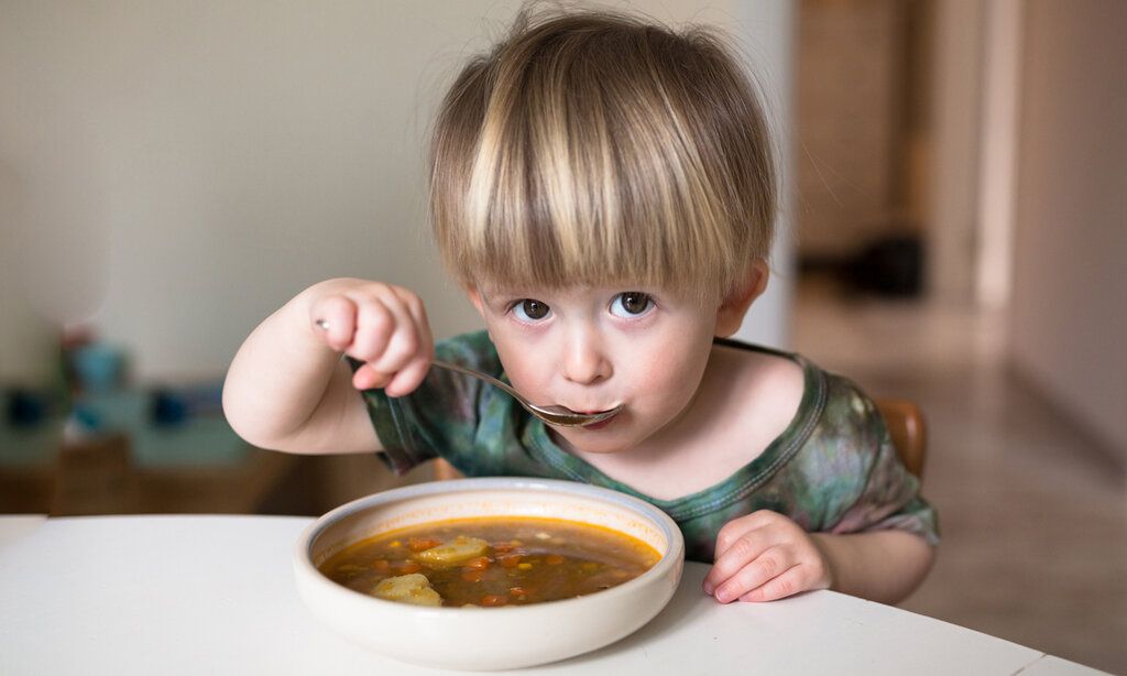 Kleines Kind sitzt an einem Tisch und isst Suppe, guckt dabei in die Kamera