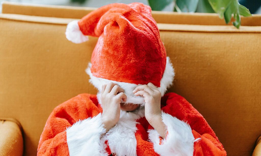 Kind mit sitzt mit einem Weihnachtsmann-Kostüm auf einem Sofa und zieht sich die Mütze übers Gesicht