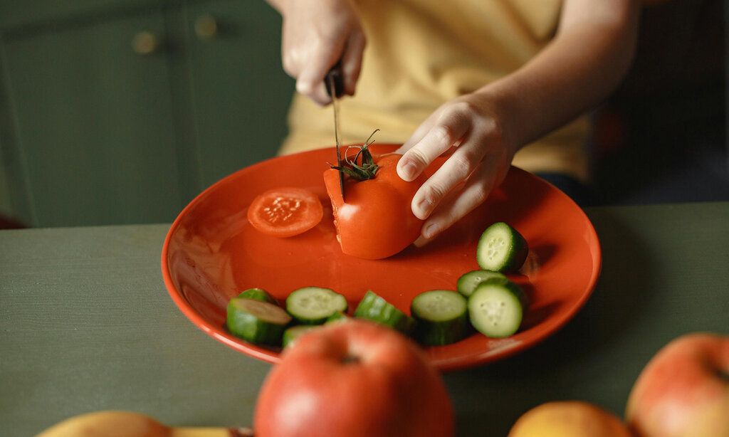 Detailaufnahme, ein Kind schneidet auf einem roten Teller eine Tomate, geschnittene Gurken liegen auf dem Teller, auf der Arbeitsplatte liegen Äpfel und Bananen