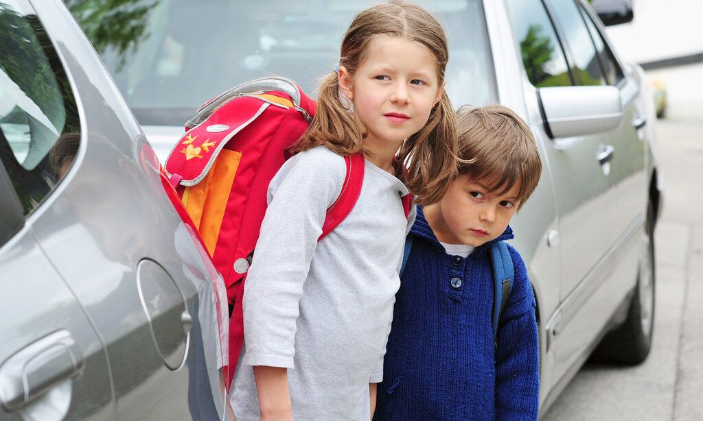 Zwei Schulkinder stehen zwischen parkenden Autos und versuchen, die Straße zu überqueren