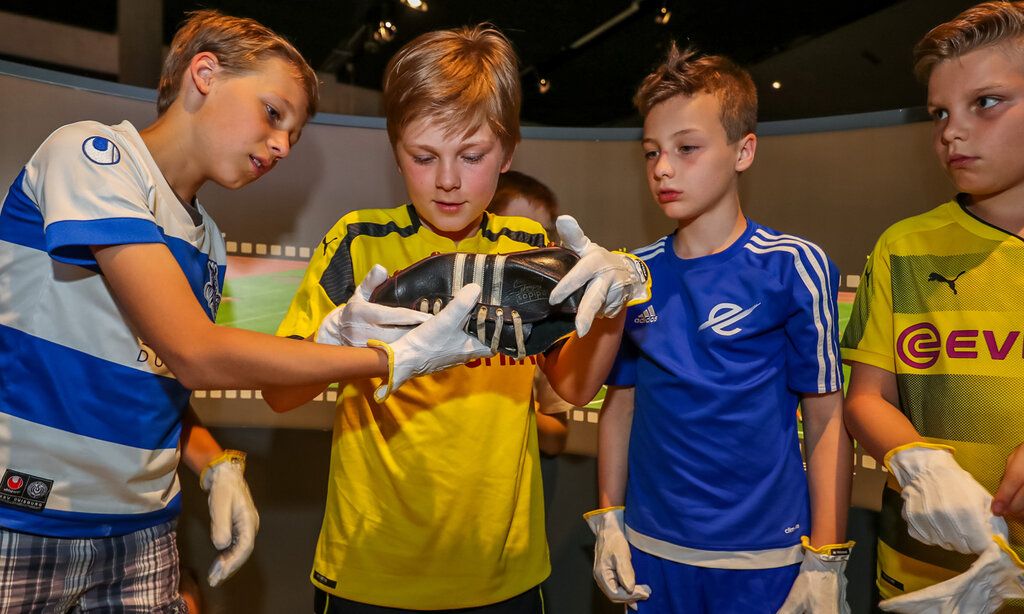 Vier Jungs in Fußballtrikots betrachten einen alten Fußballschuh, den einer der Jungs in der Hand hält