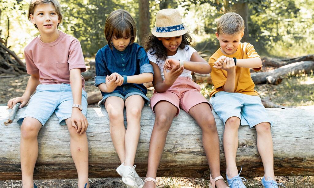 Vier Kinder sitzen auf Baumstamm und gucken auf ihre Armbanduhren