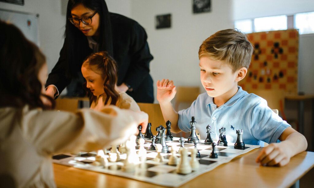 zwei Kinder spielen Schach, im Hintergrund eine Frau, die mit einem Mädchen spricht