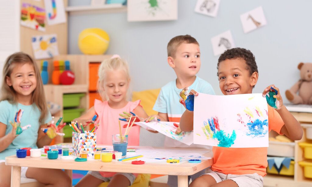 4 Kinder sitzen an einem Kindertisch, bemalenihre Hände und machen Handabdrücke auf Papier, ein Junge hält sein Bild in die Kamera