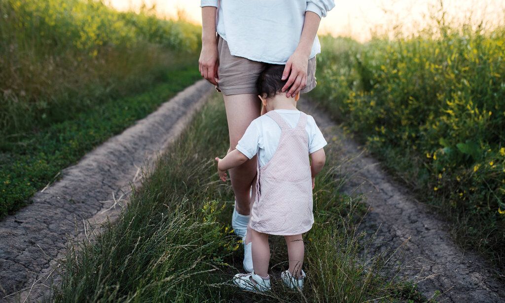 Kleinkind steht an den Beinen der Mutter, umfasst die Beine, beide stehen auf einem Feldweg, umgeben von Feldern