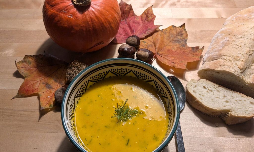 Eine Schale mit Kürbissuppe, daneben liegt ein Löffel, aufgeschnittenes Brot, Herbstlaub, Kastanien und ein Hokkaido-Kürbis