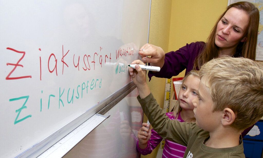 Eine Lehrerin und zwei Kinder stehen an einem White Board, ein Junge schreibt etwas auf das Board