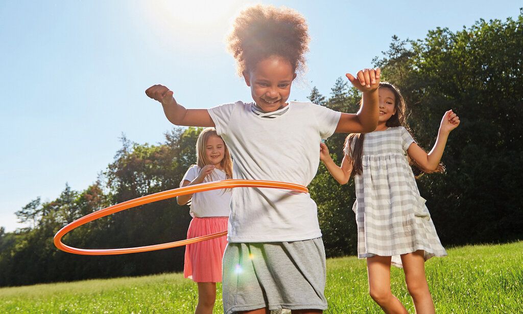 Drei Mädchen auf einer Sommerwiese, eines lässt den Hula-Hoop-Reifen kreisen