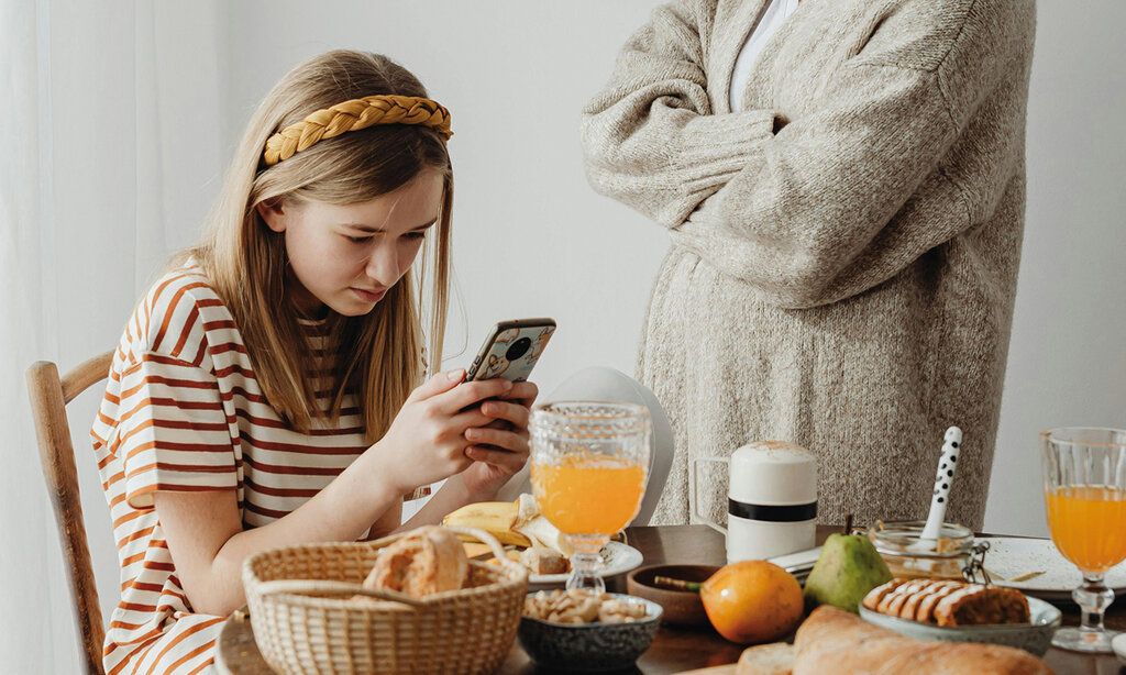 Mädchen sitzt am Frühstückstisch und daddelt, die Mutter steht mit verschränkten Armen daneben