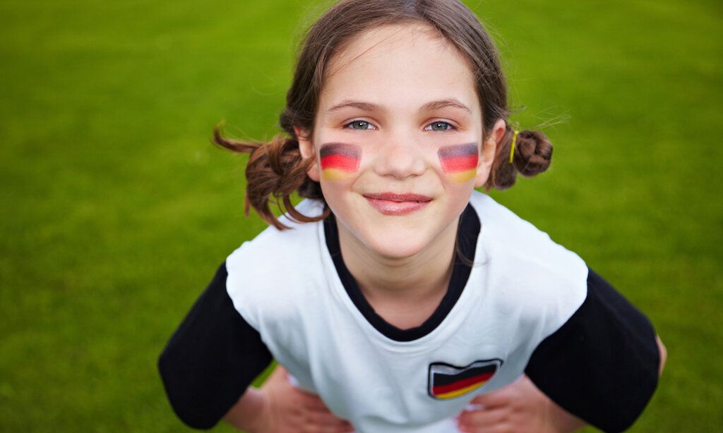 Ein Mädchen steht auf grünem Rasen und guckt in die Kamera, auf den Wangen ist die Deutschlandflagge geschminkt, sie trägt ein Deutschland-Trikot