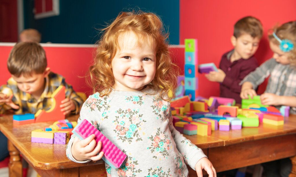 Kleines Mädchen mit einem bunten Bauklotz in Kita, im Hintergrund andere Kinder die an einem Tisch mit Bauklötzen spielen