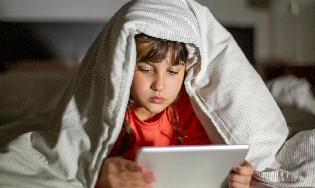 Mädchen liegt im Bett und schaut auf sein Tablet, die Betdecke liegt auf dem Kopf