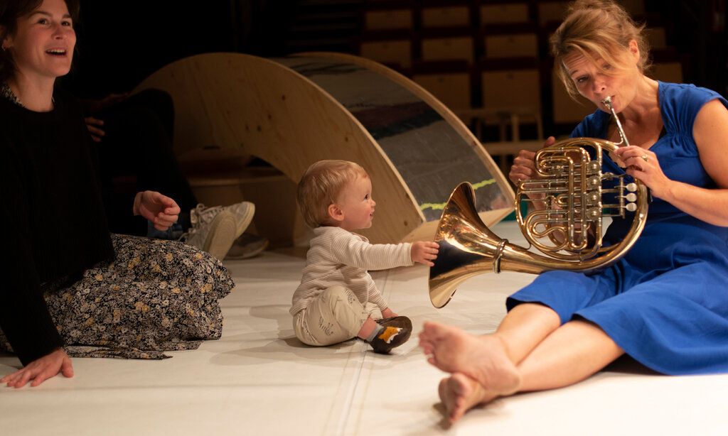 Frau im blauen Kleid spielt Horn, ein Baby guckt zu und fasst das Instrument an, beide sitzen auf der Erde, eine andere Frau sitz dabei und lacht