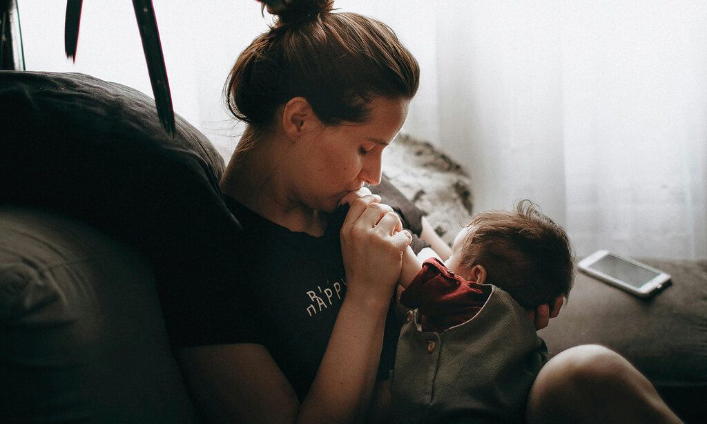 eine Mutter sitzt mit ihrem Baby auf einem Sofaund küsst dessen Händchen