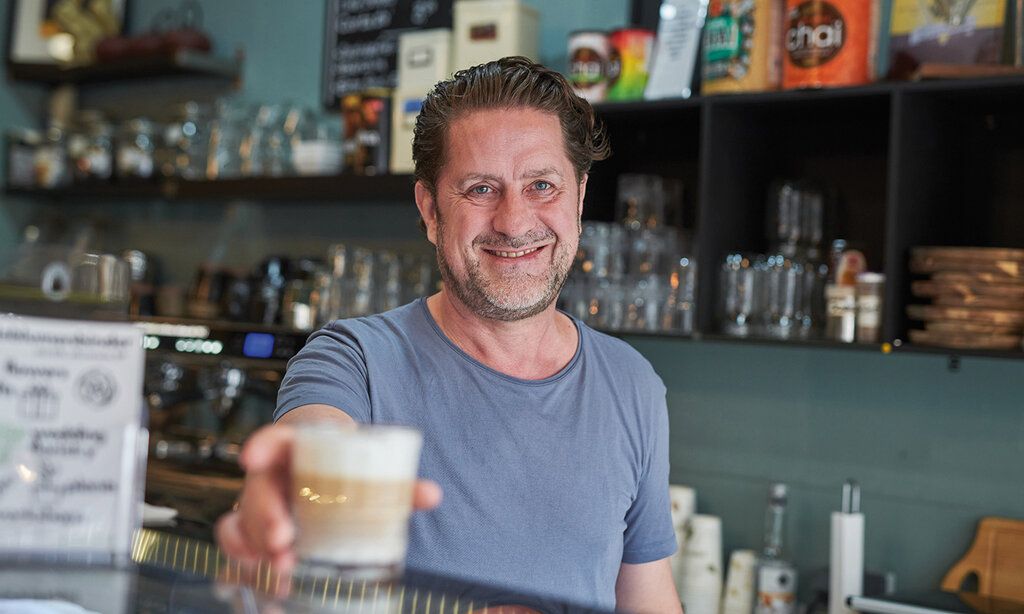 Mann lächelt in die Kamera und stellt einen Kaffee auf die Theke des Cafés Genußstation