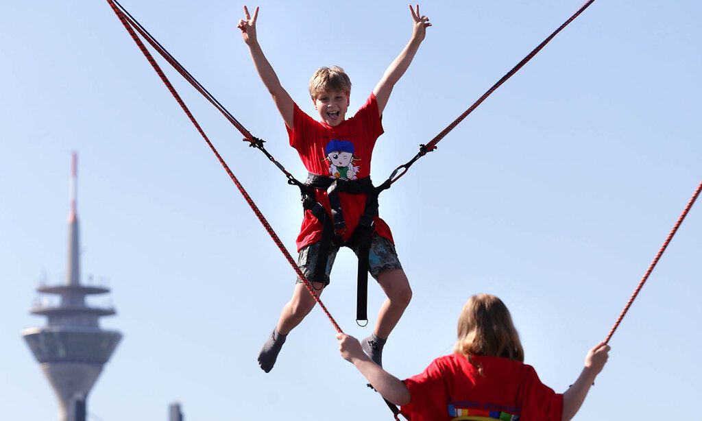 Ein Junge hüpft auf einem Trampolin und macht dabei Victory-Zeichen, im Hintergrund der Rheinturm, davor Mädchen von hinten