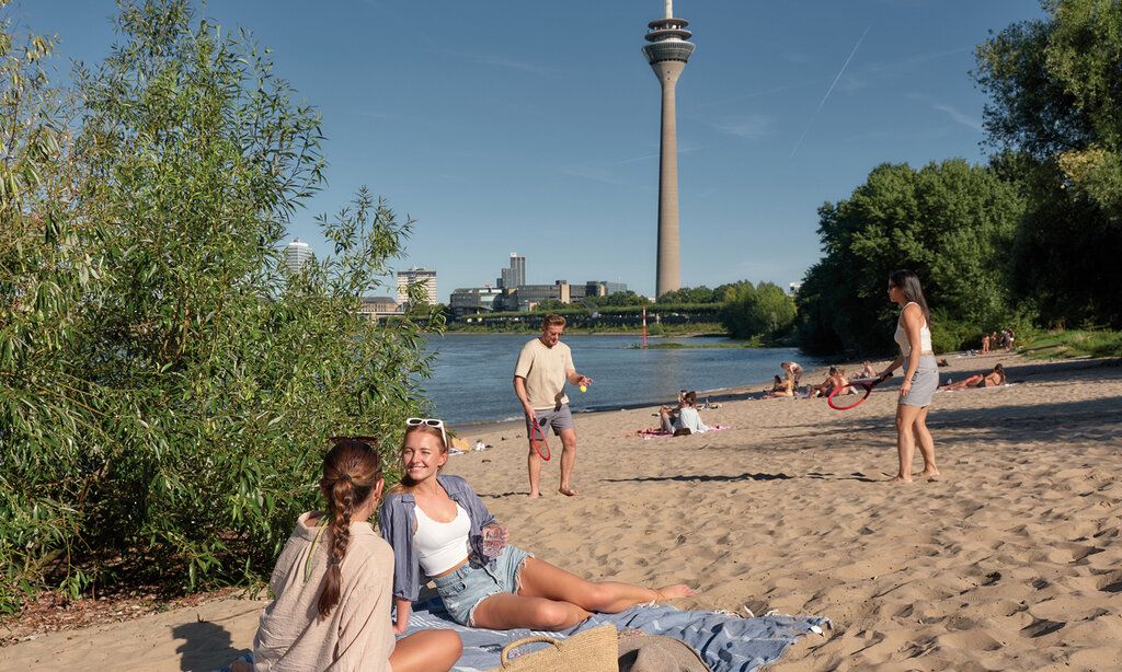 Paradiesstrand Düsseldorf, zwei Frauen sitzen im Sand, ein Man und eine Frau spielen Beachball, im Hintergrund der Fernsehturm