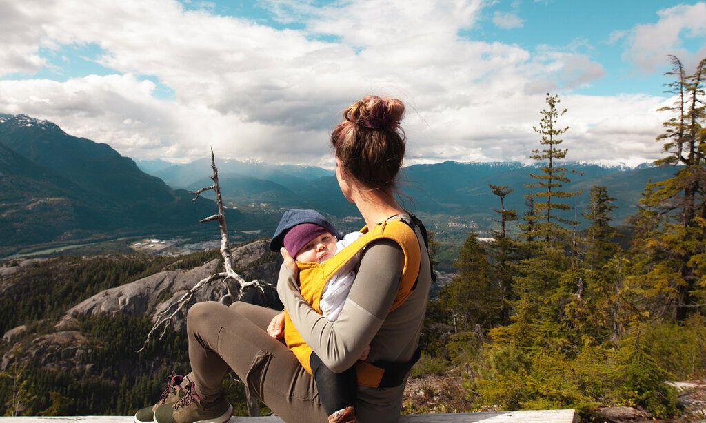 Frau mit Baby im Tragegurt, blickt auf Bergkulisse