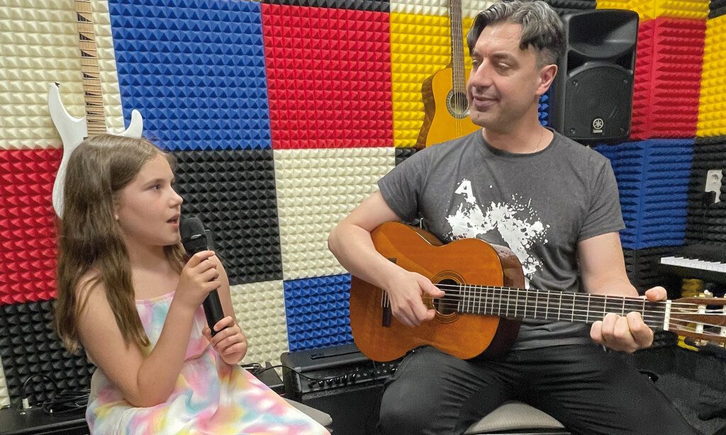 Ein Mädchen sitzt mit seinem Musiklehrer zusammen. Das Mädchen singt in ein Mikrofon, der Lehrer spielt Gitarre. Im Hintergrund sieht man, dass der Raum schallgedämpft ist, die Wände sind mit blauen, roten, schwarzen ud weißen Platten verkleidet, Musikinstrumente, Verstärker und Boxen sind zu sehen