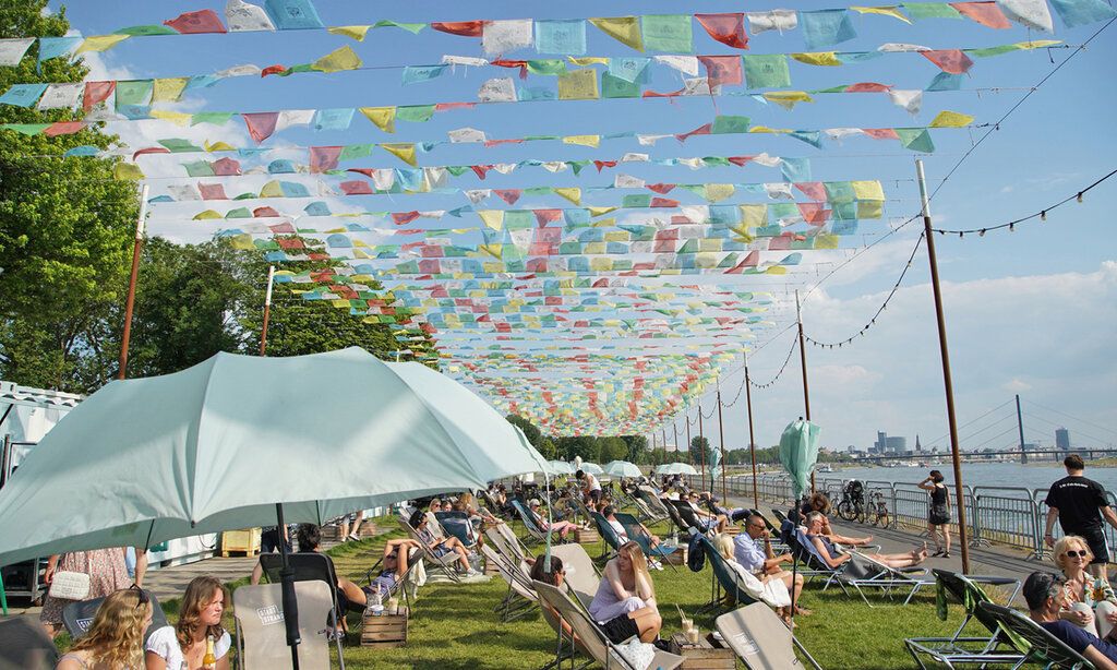 Stadtstrand Robert Lehr Ufer, Leute liegen auf Liegestühlen auf der Wiese, tibetische Gebetsfahnen flattern im Wind, Rhein mit Blick auf Kniebrücke im Hintergrund