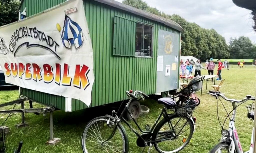 Bauwagen mit einem Banner, auf dem steht: Kinderstadt Superbilk, auf einer Wiese, im Hintergrund Leute, im Vordergrund Fahrräder