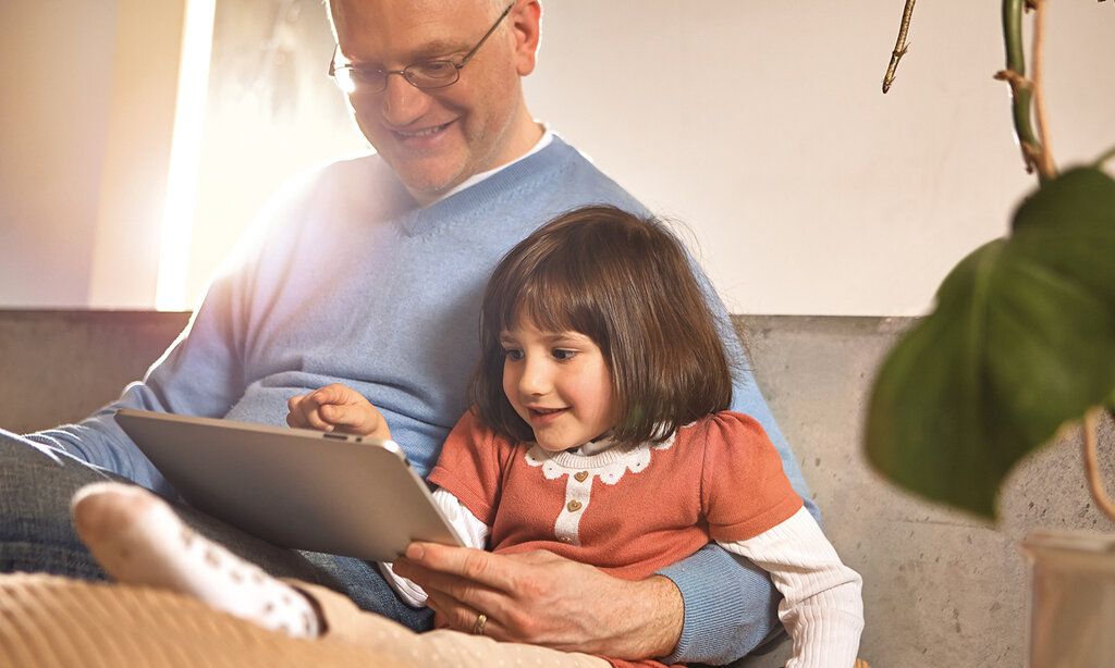 Opa sitzt mit Enkelin zusammen und beide schauen aufs Tablet.