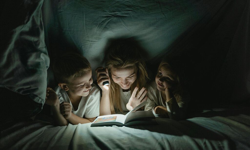 Mutter liest mit zwei Kindern unter der Bettdecke mit der Taschenlampe ein Buch