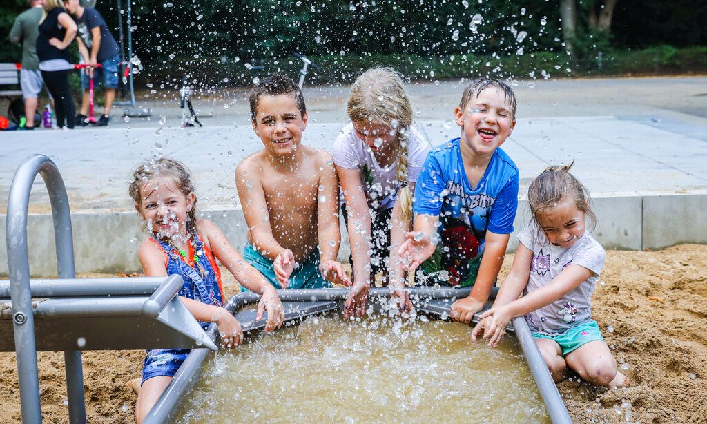 5 Kinder spielen an einer Wasseranlage auf einem Spielplatz, Wasser spritzt, die Kinder lachen