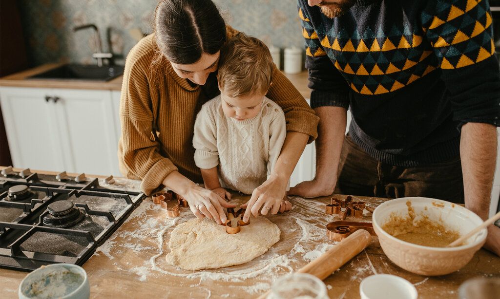 Kleinkind mit Eltern in der Küche, die Mutter hilft ihm dabei, Plätzchen aus Teig auszustechen