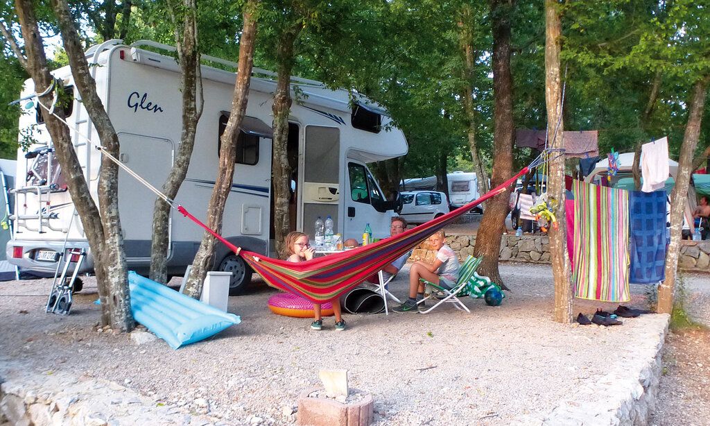 Ein Wohnmobil steht auf einem Campingplatz, zwischen Bäumen ist eine Hängematte gespannt, Familie sitzt vor Wohnmobil