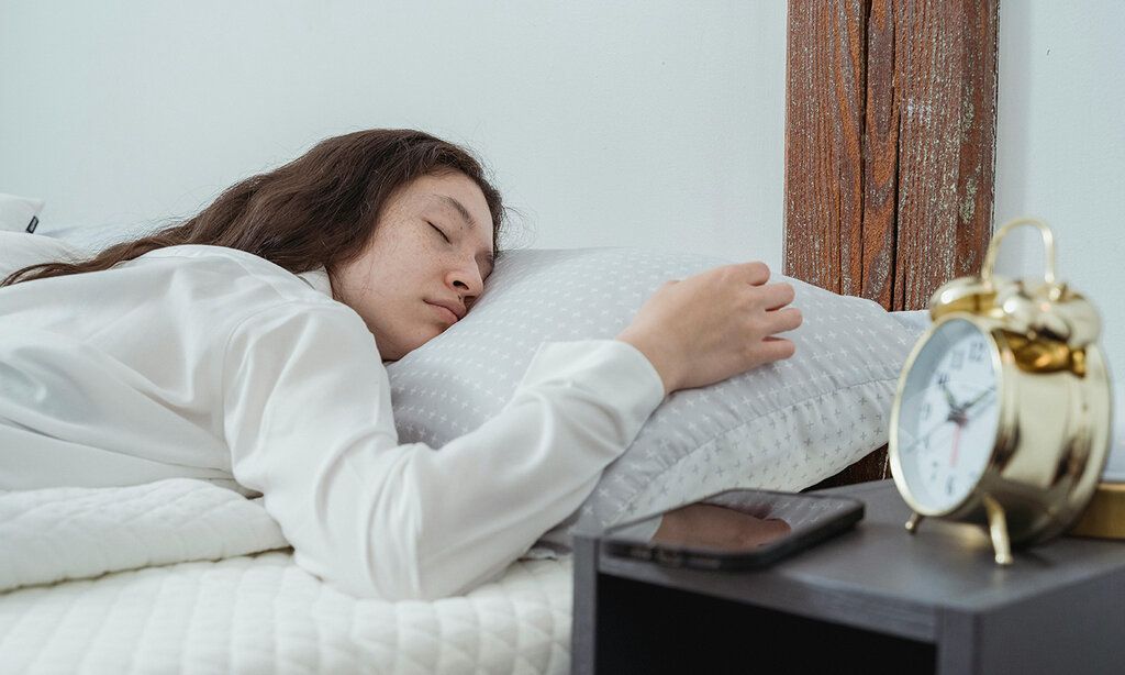 Frau im weissen Hemd liegt auf weissem Bett mit einem goldenen Wecker an der Seite